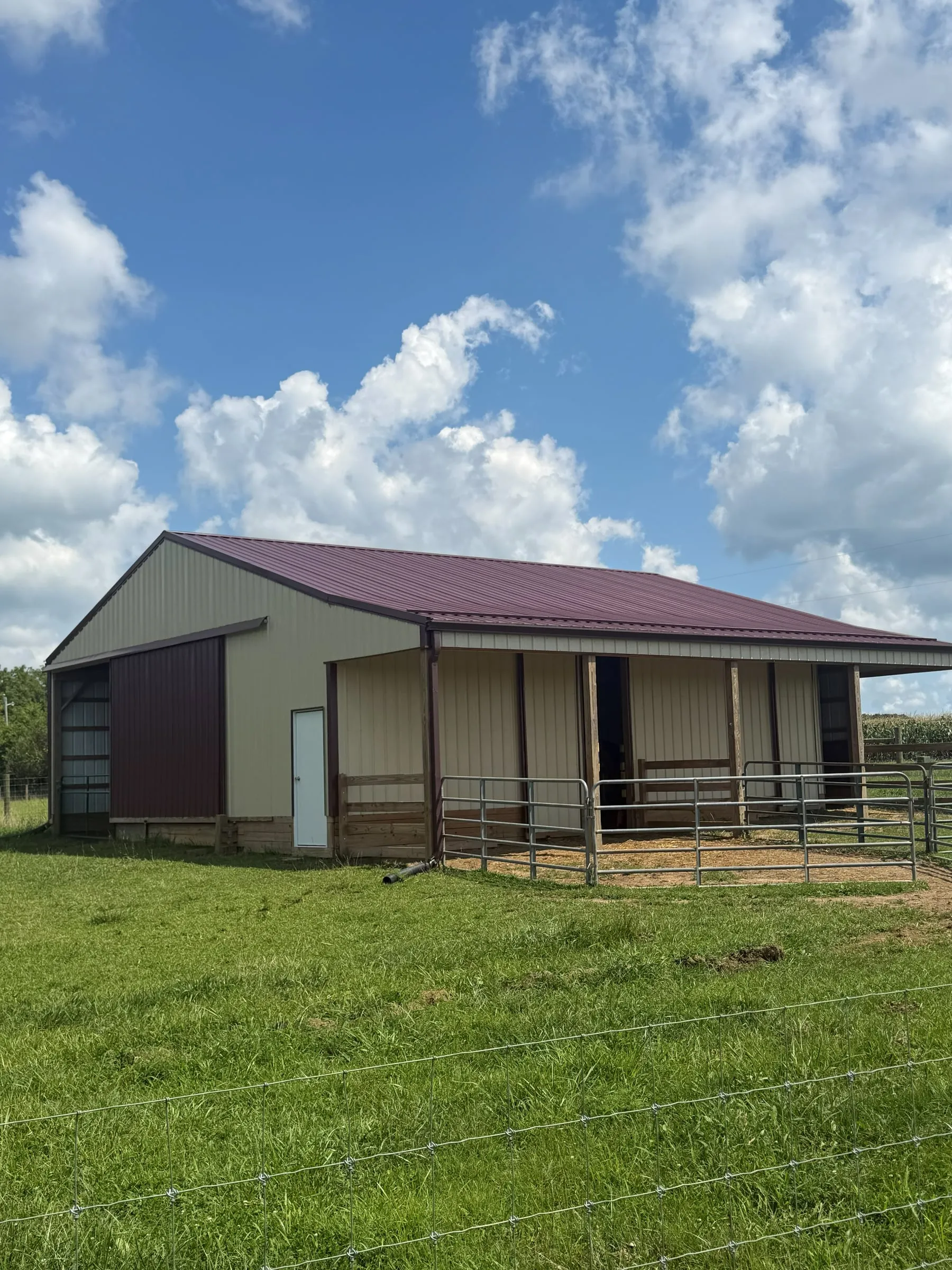 Maroon-roofed farm building with a white gable end and an open lean-to over a board fence, in an open pasture