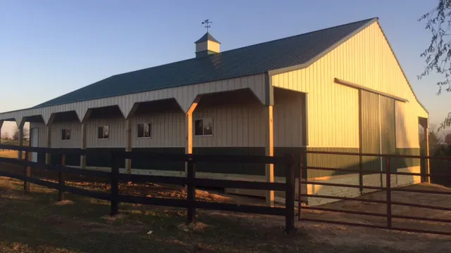 Tan horse barn with a dark roof, open stall fronts on timber posts, a cupola and a fenced paddock
