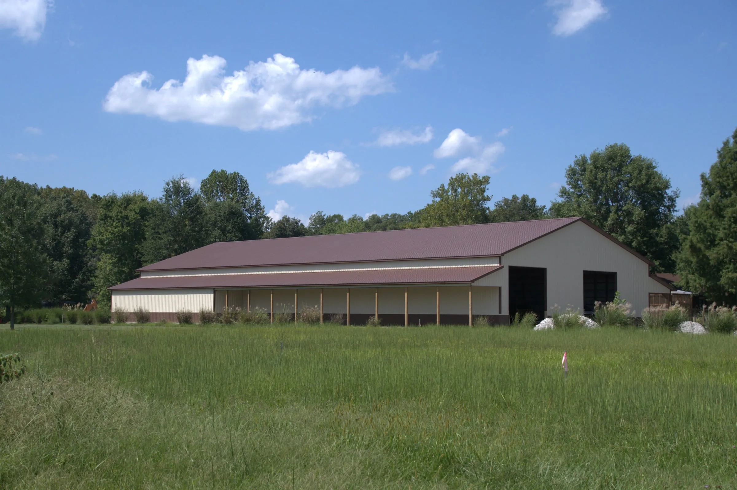 Long tan building with a maroon roof and a row of open bays along the side, set in a mowed field