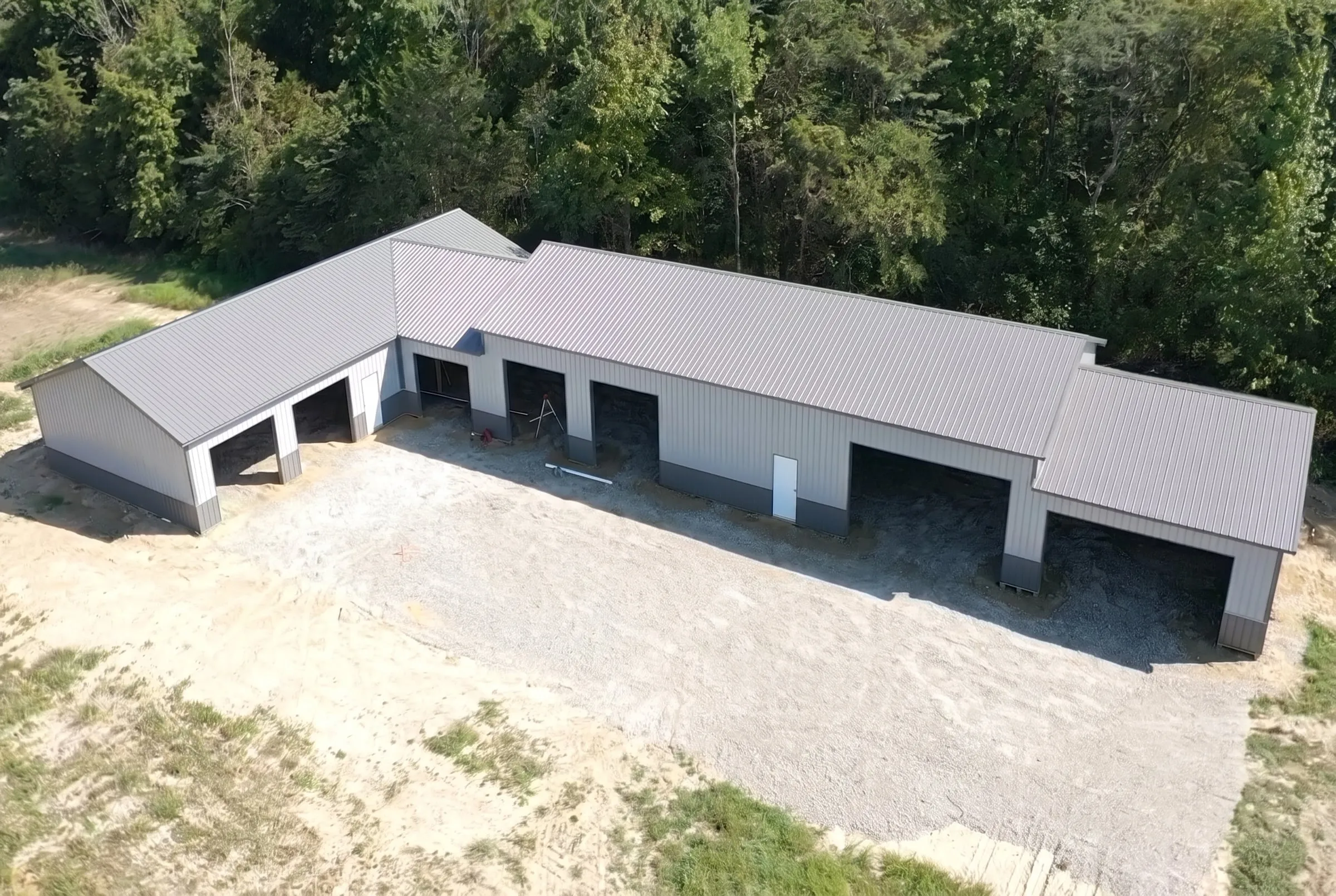 Aerial view of a long gray L-shaped farm building with open bays, on a fresh gravel pad at the edge of the woods