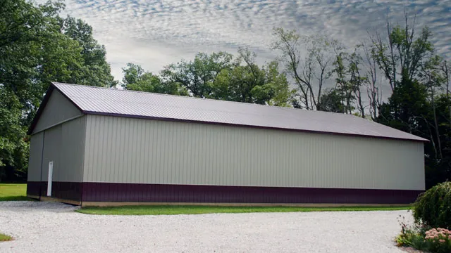 Cream barn with a maroon roof and maroon wainscot standing on gravel