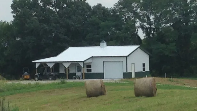 White barn with green trim, an open equipment bay and round hay bales in front