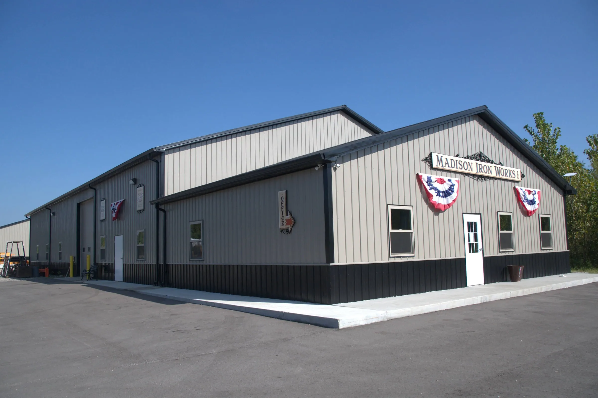 Two-part commercial building in gray with charcoal wainscot, a front office with a sign and flag bunting, and a taller shop behind