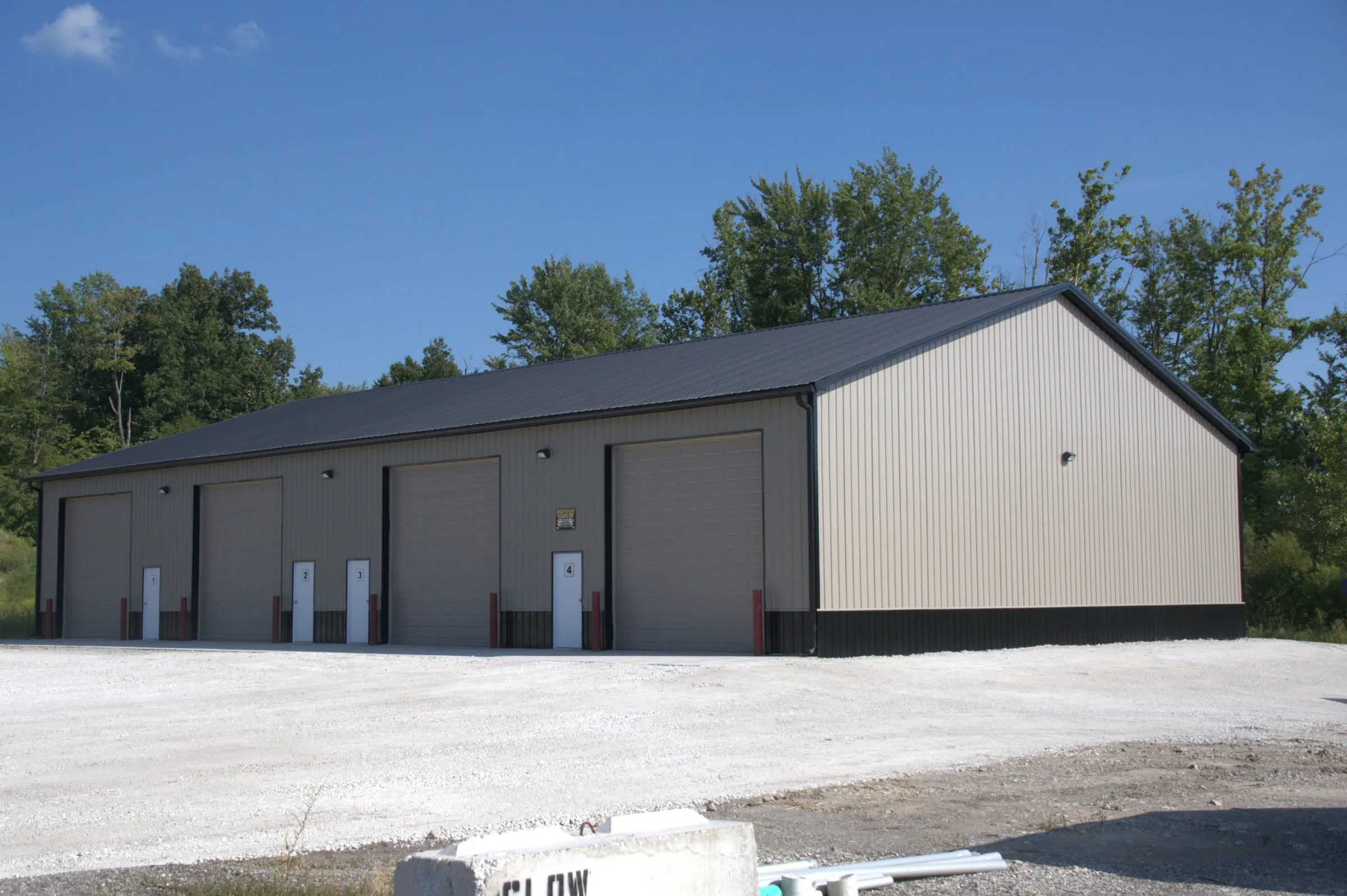 Long white commercial building with a black roof and a row of overhead doors along the side, on a new concrete lot
