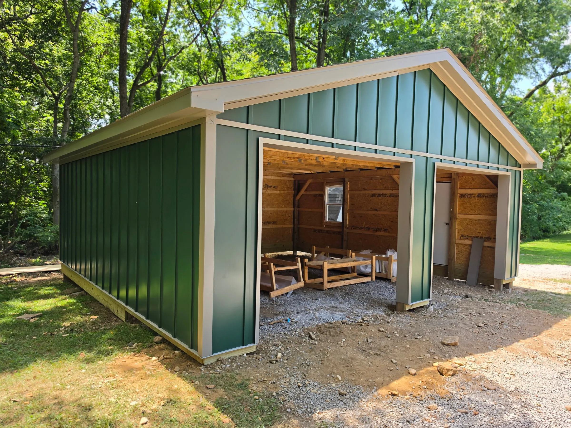 Green board and batten shed with white trim and open equipment bays, in the trees