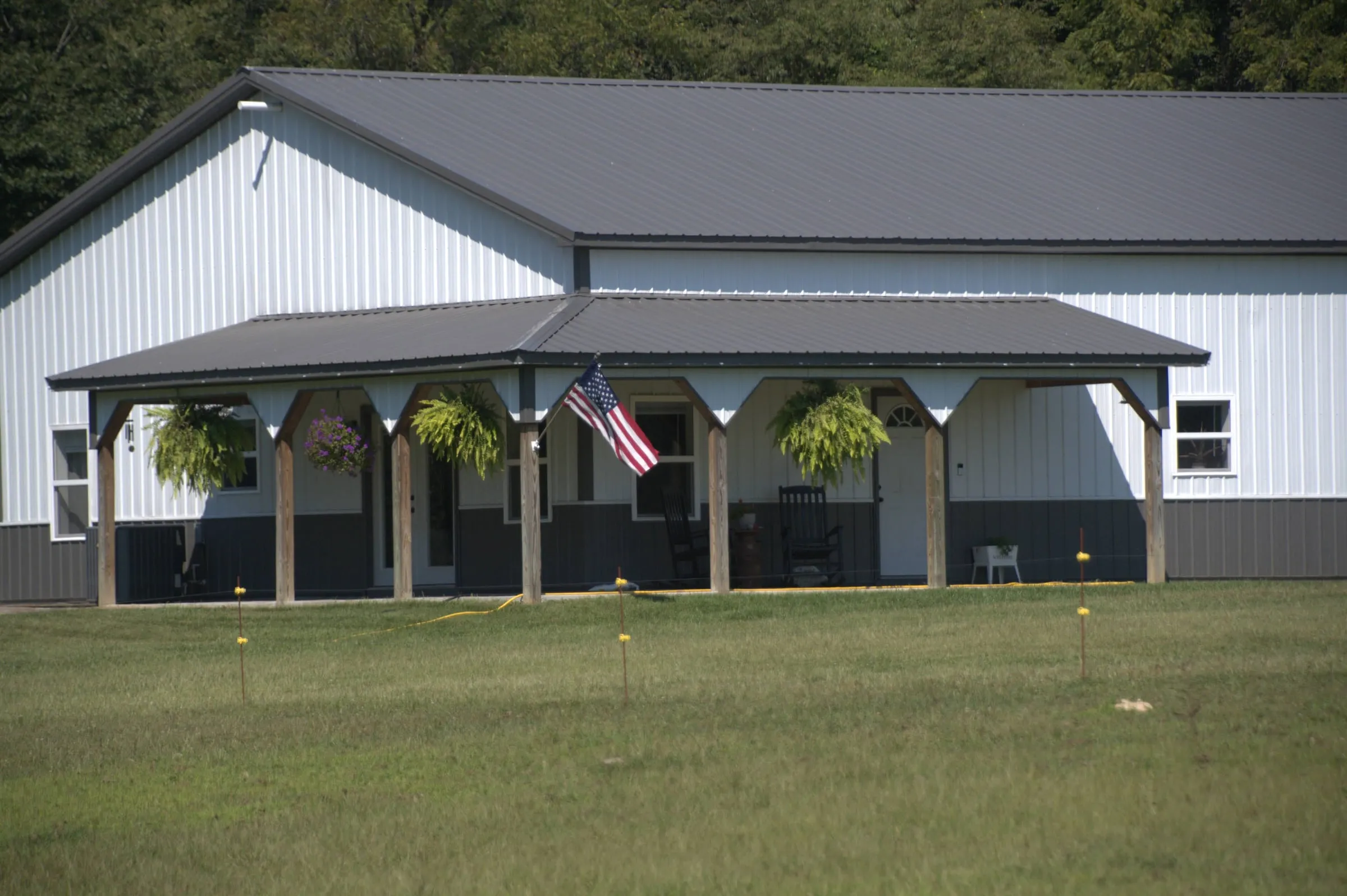 White building with a gray roof, charcoal wainscot and a covered porch across the front with hanging baskets and a flag