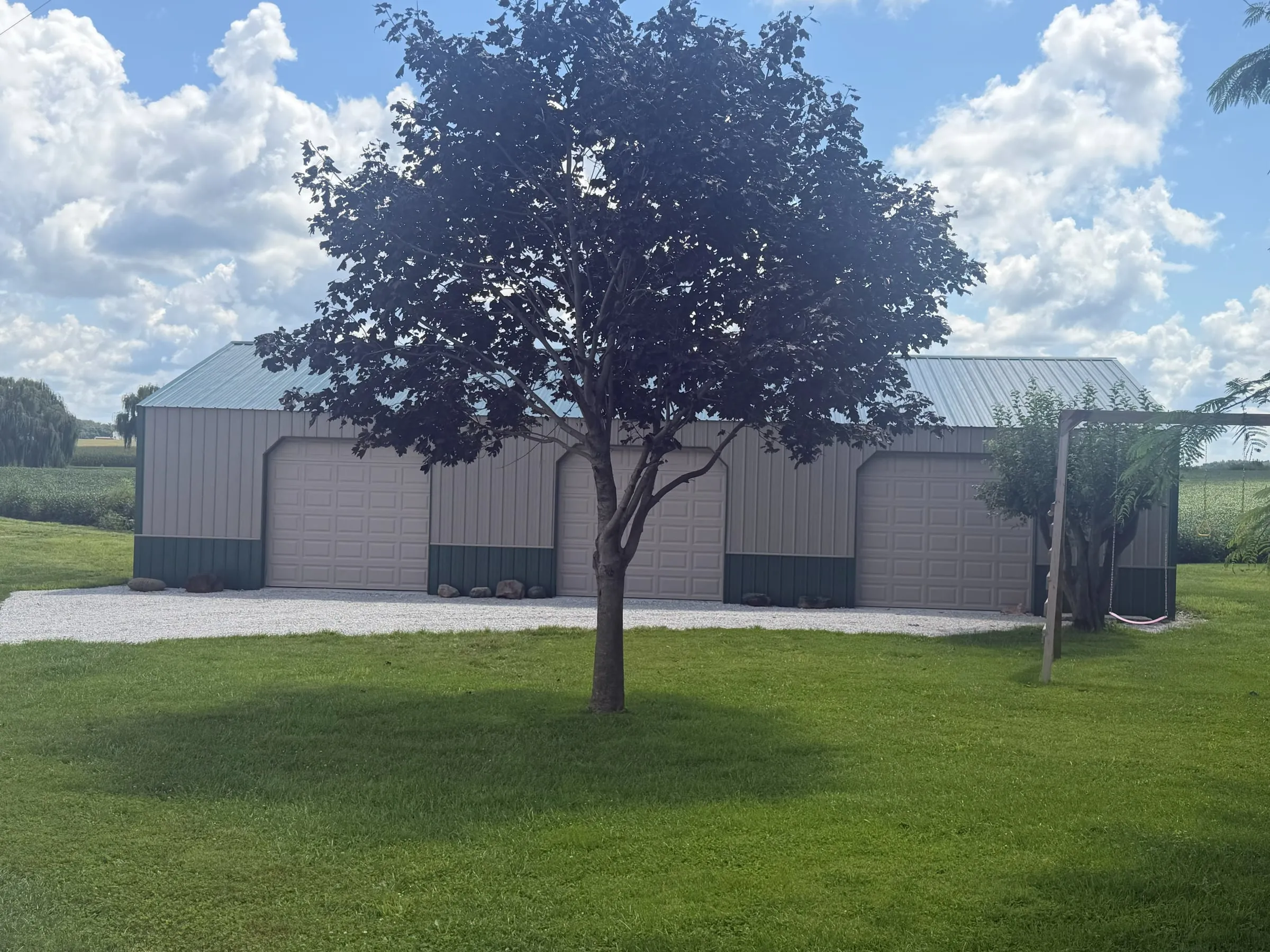 Long tan building with a gray roof and three bays, under a large shade tree on a mowed lot