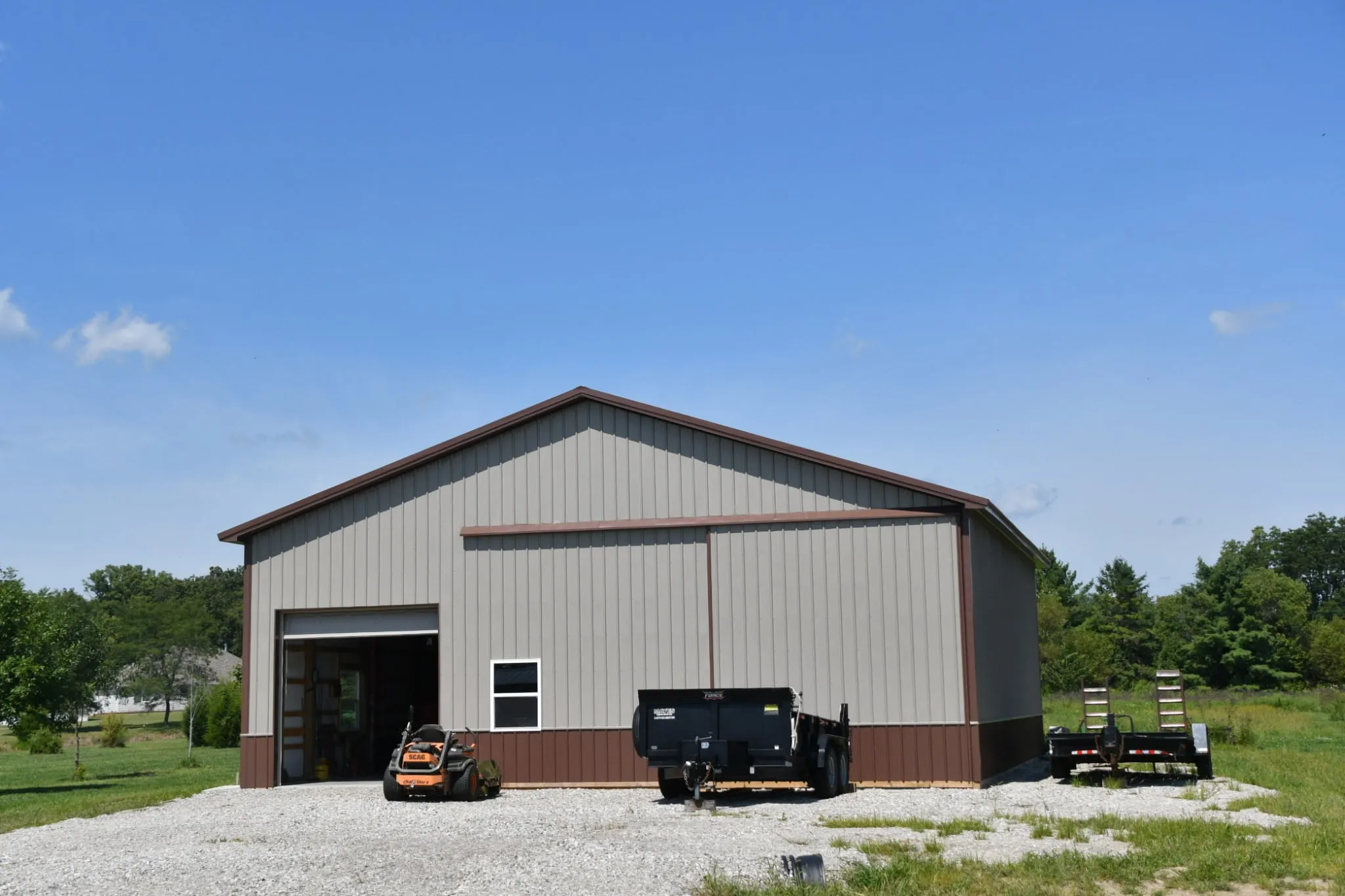 Tall gray shop with brown trim and wainscot, a wide overhead door and a walk door, equipment parked outside
