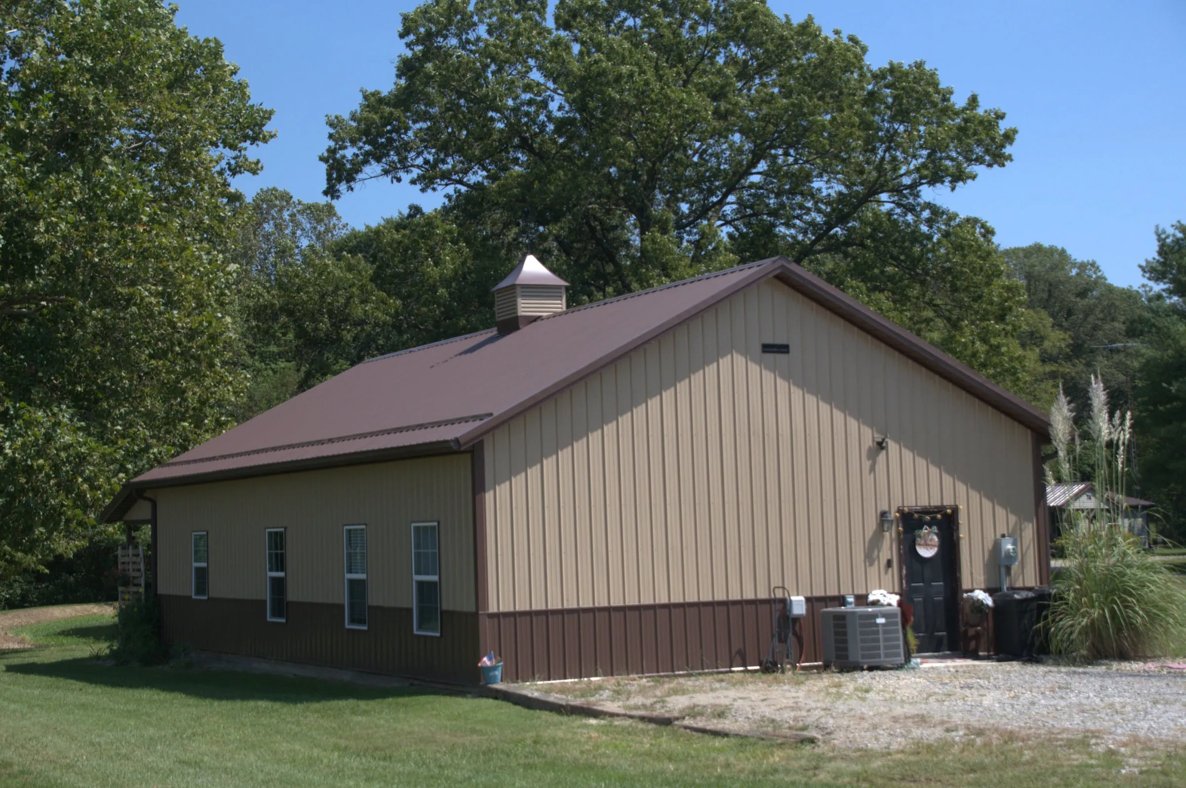 Tan shop with a maroon roof and brown wainscot, a cupola, windows and a walk door, on a gravel drive