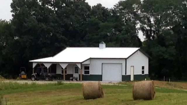 White building with an open equipment bay and round hay bales in the field in front