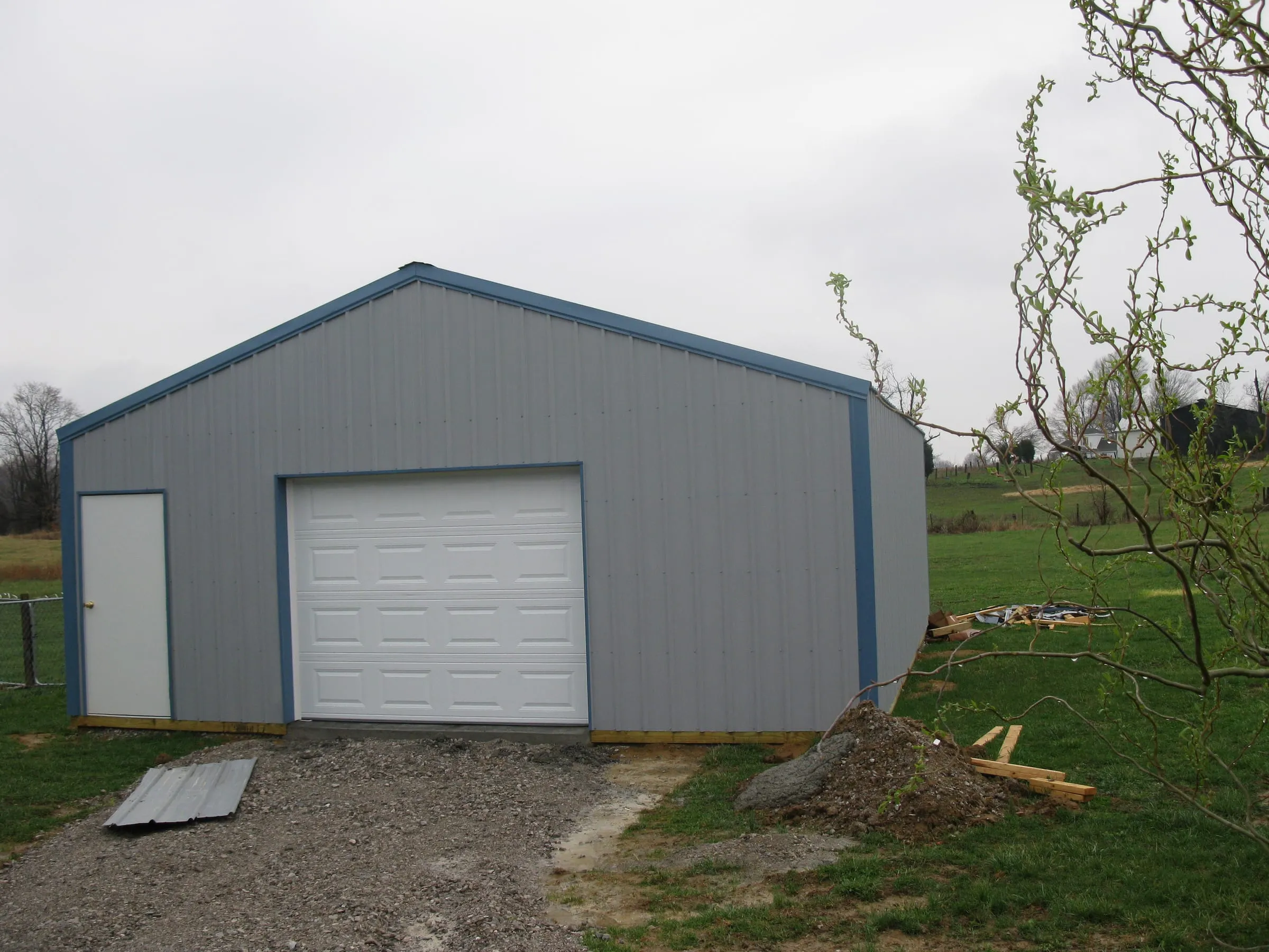Gray shop with blue trim and a white overhead door beside a walk door, on fresh gravel