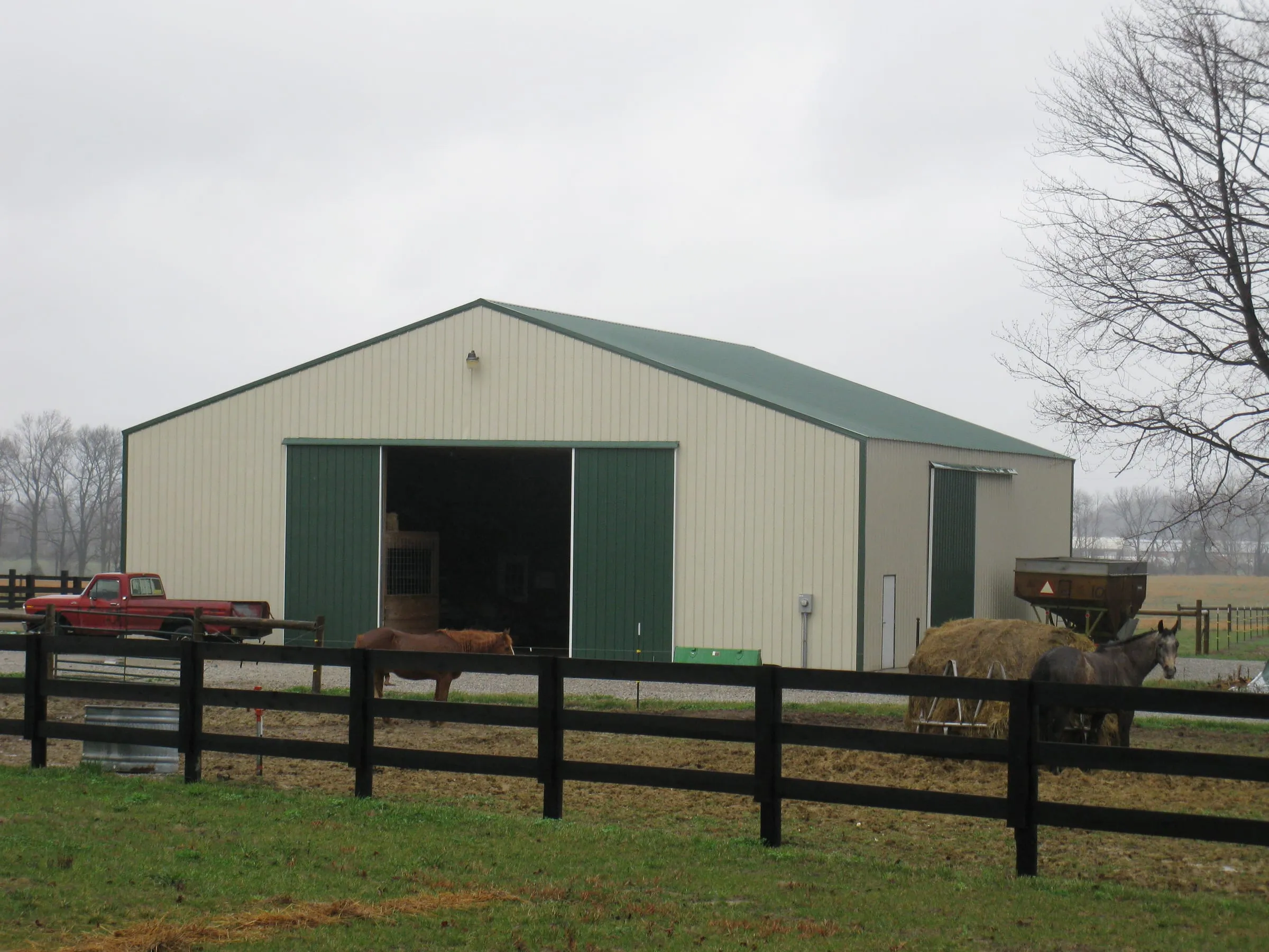 Cream barn with a green roof and trim, an open bay holding equipment, black board fence in front