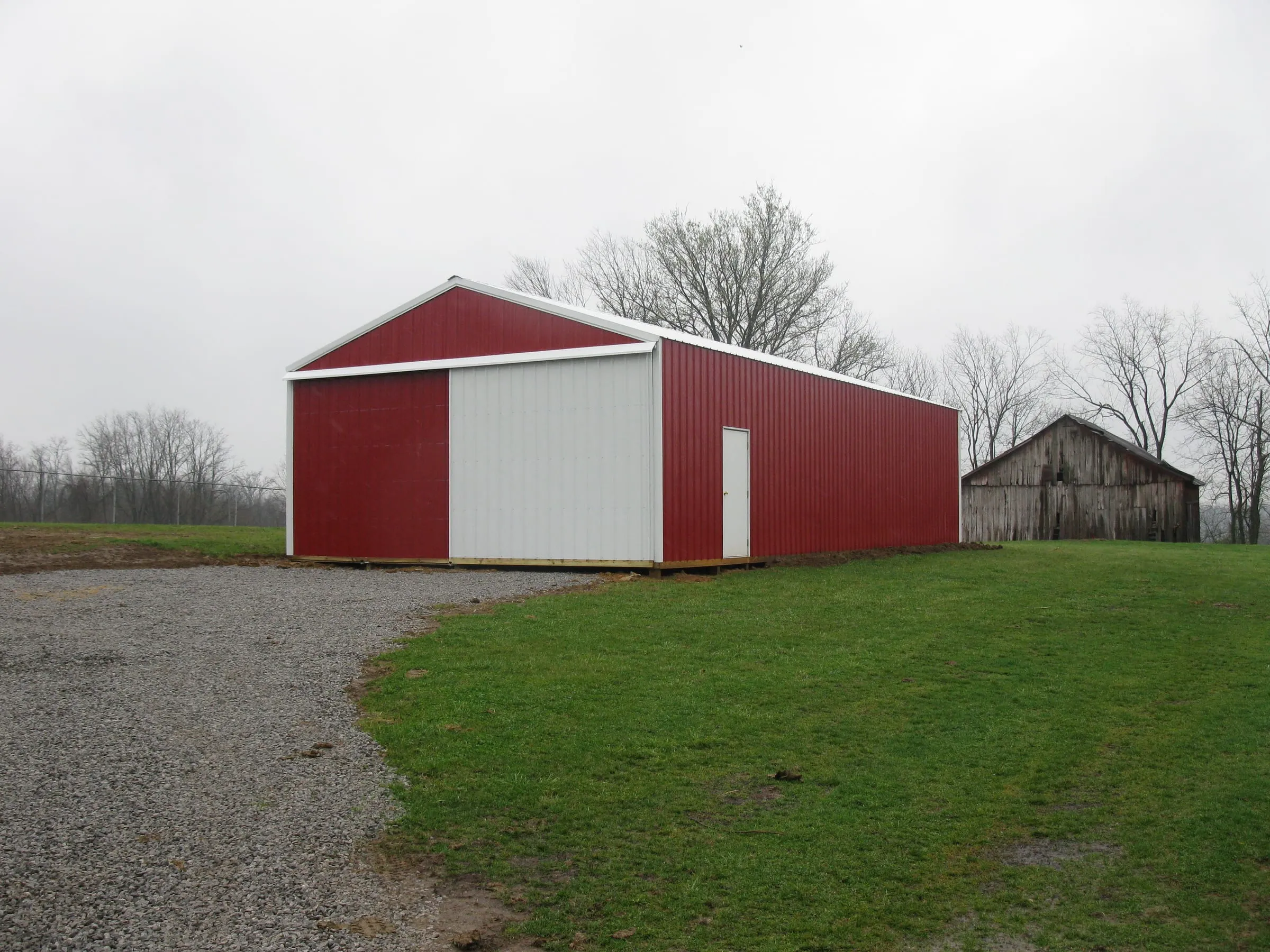 Red barn with a white gable and a dark roof on gravel, an older barn standing behind it