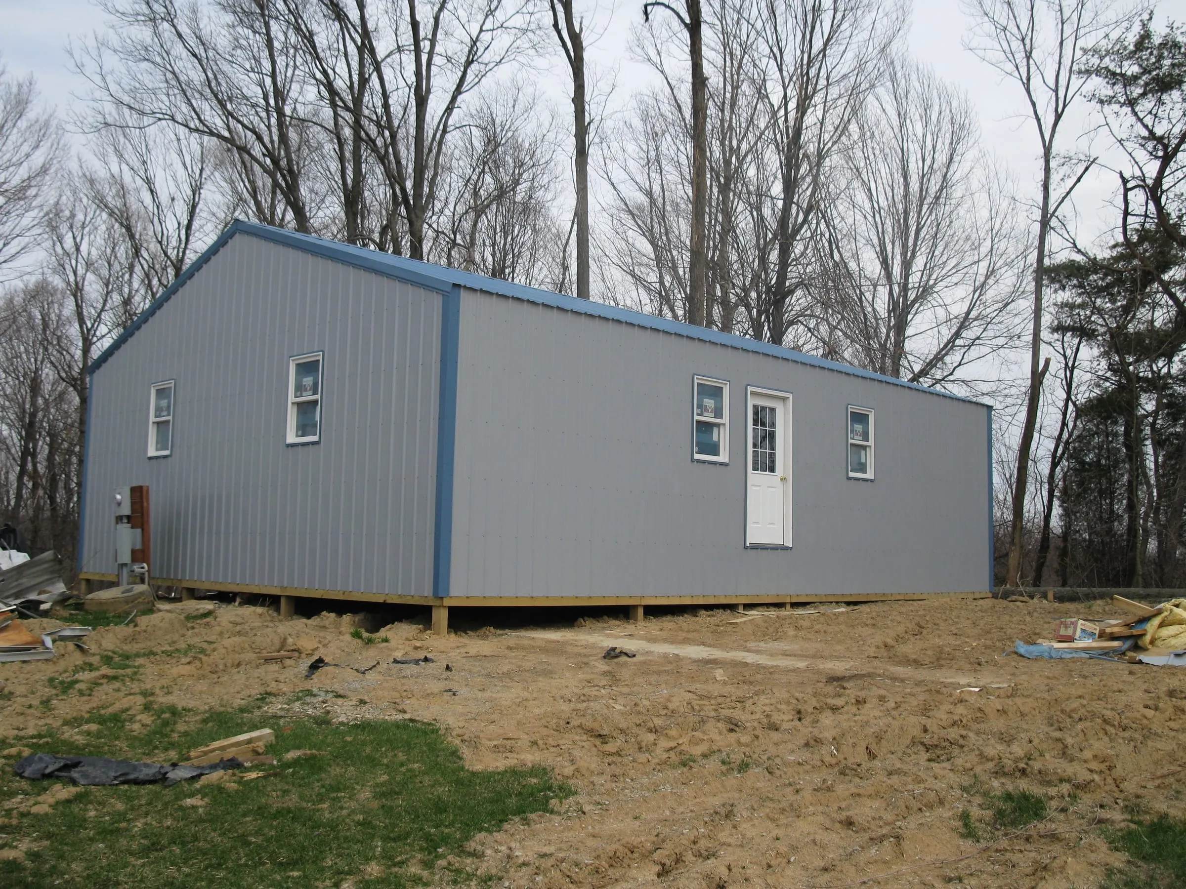 Gray-blue building with white trim, three windows and a white walk door, standing on fresh dirt