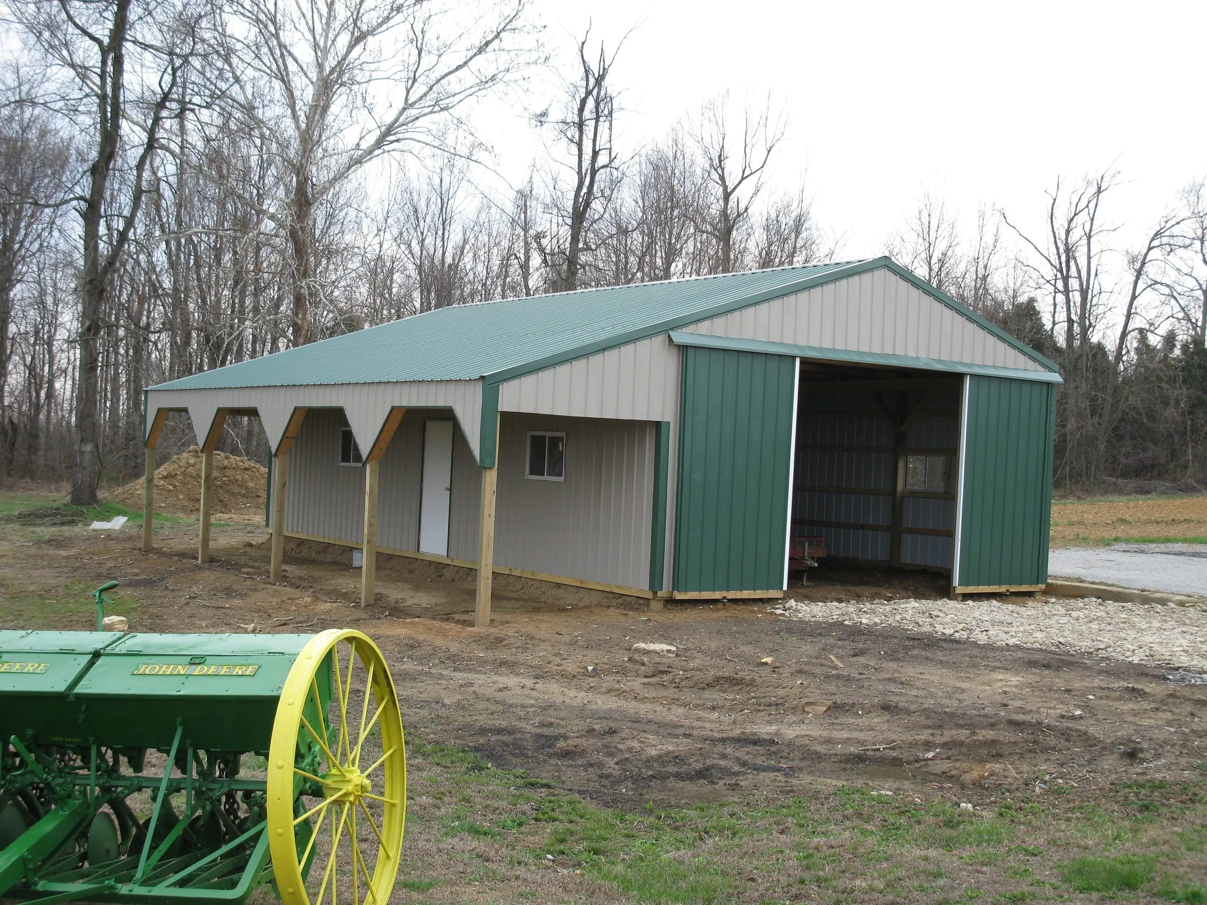 Cream barn with a green roof and timber posts along an open side, a grain drill in the foreground