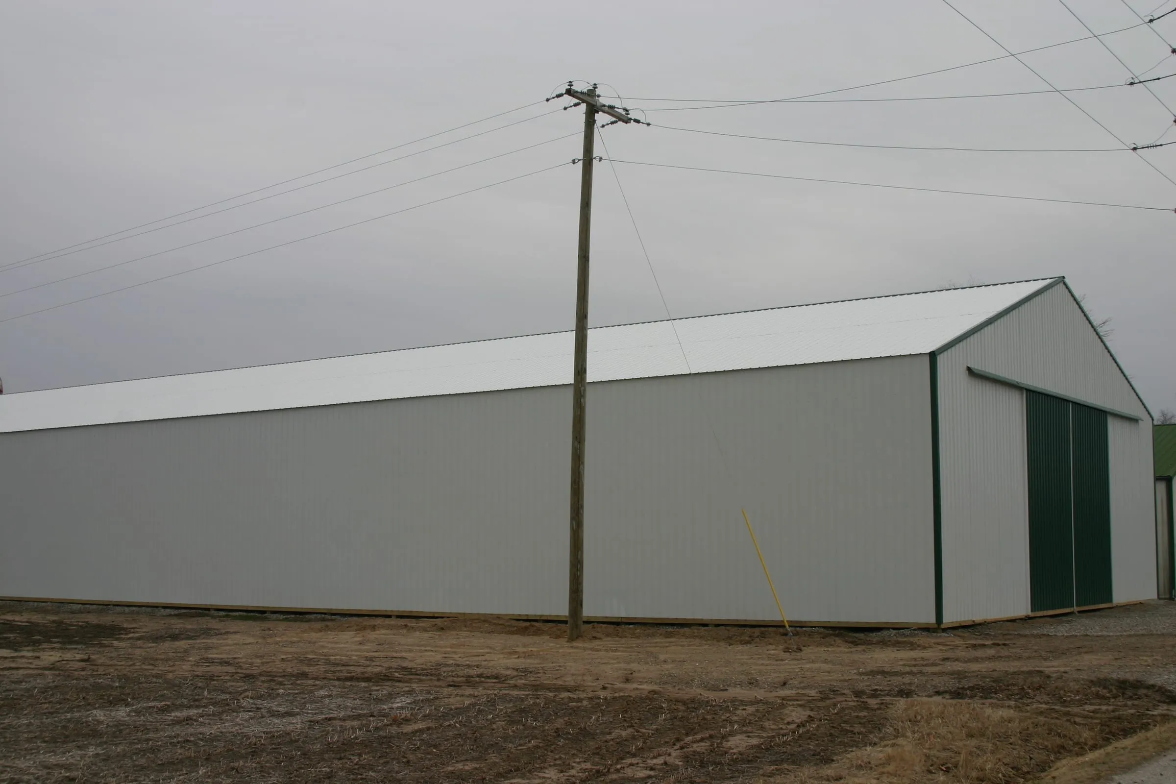 Long white machine shed with a green sliding door, power line and worked ground in front