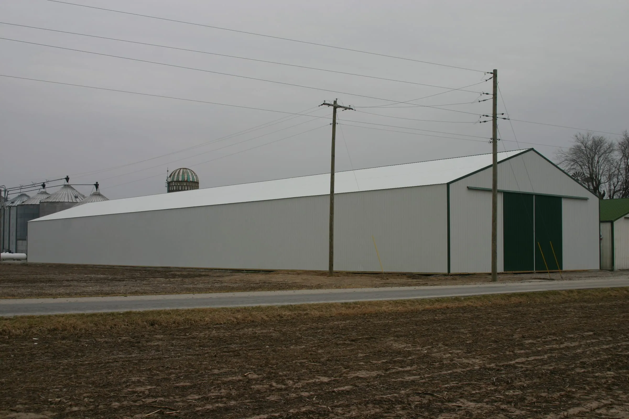 Long white machine shed with a green sliding door and grain bins at the far end