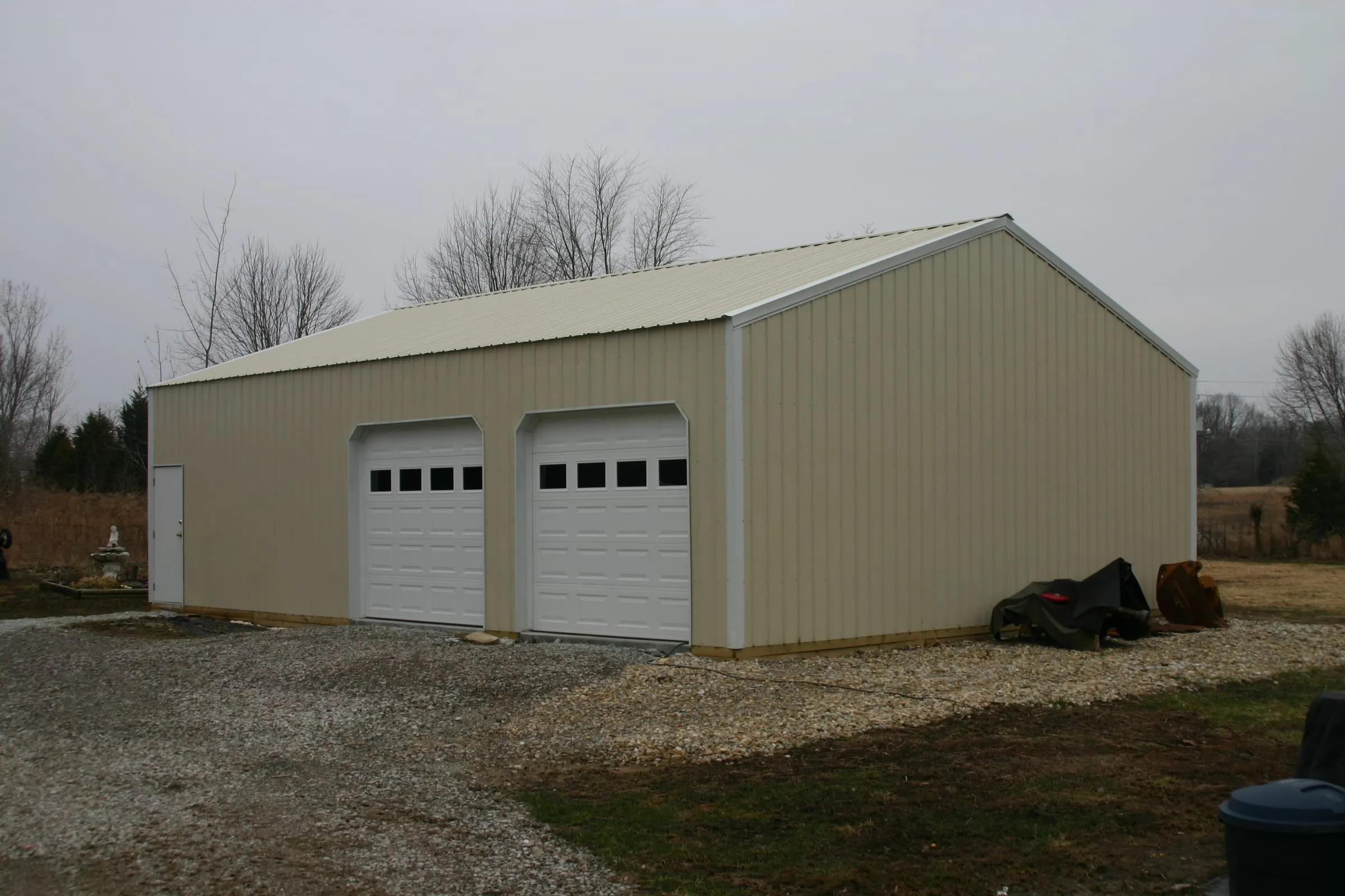 Tan shop with two white overhead doors standing on a gravel pad