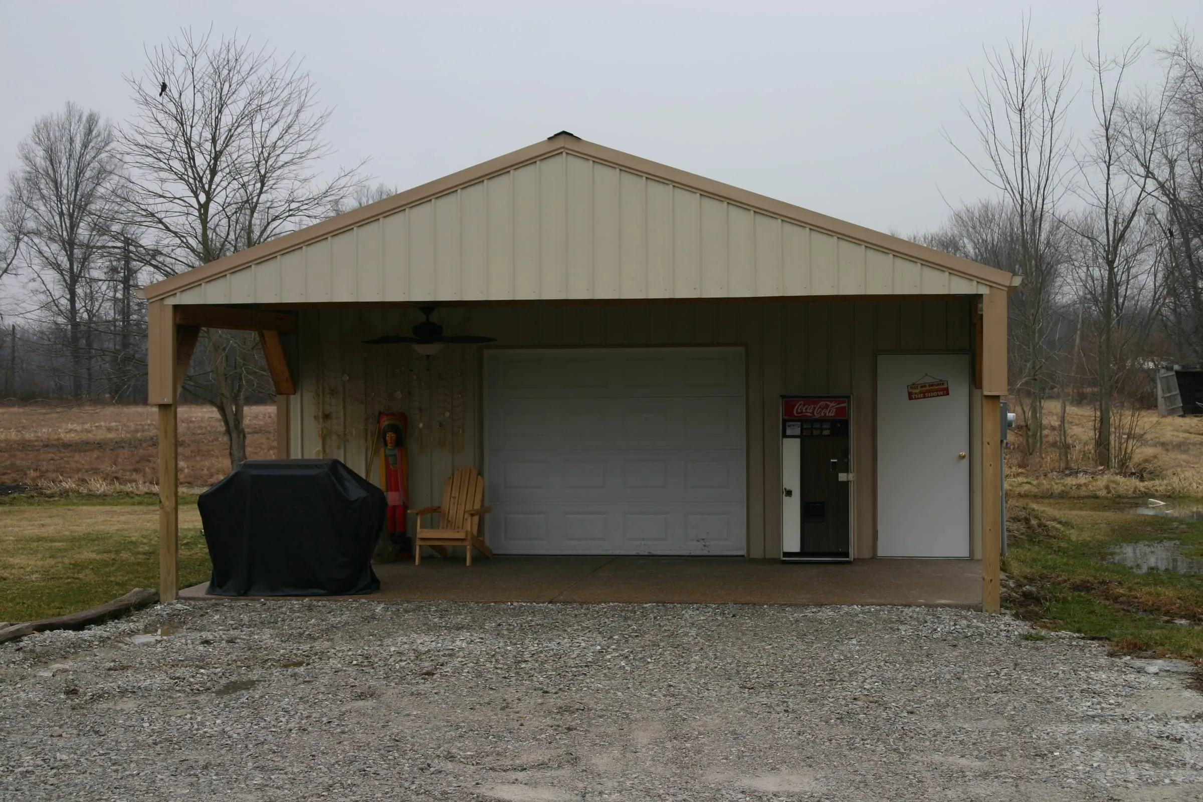 Cream building with a gable porch on timber posts over the overhead door, grill and chair under it