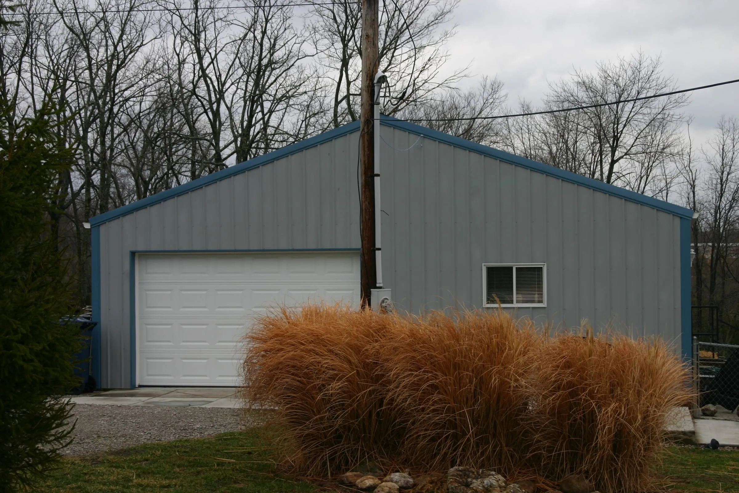 Gray building with blue trim, a white overhead door and a window, ornamental grass in front