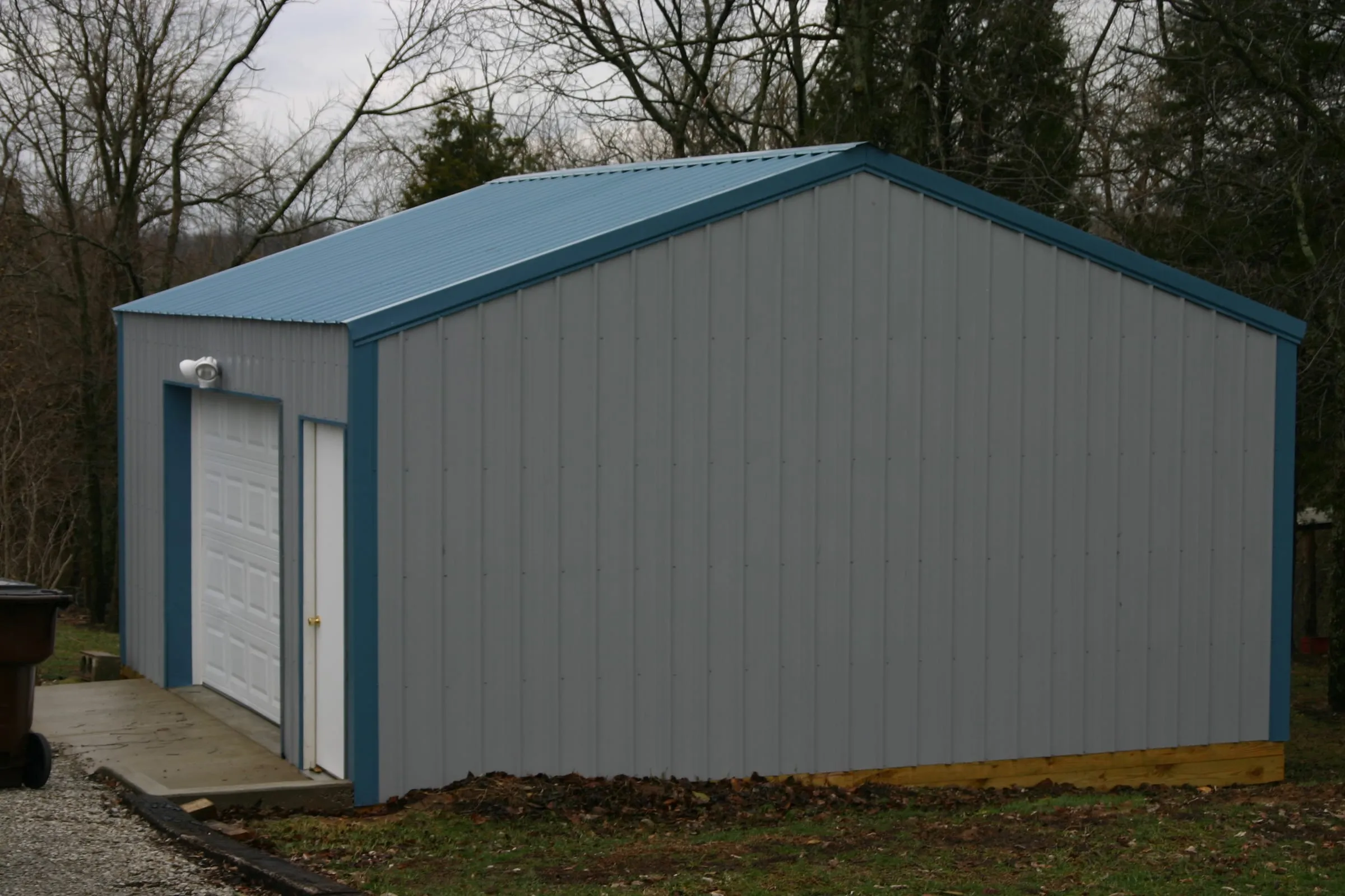Gray building with a blue roof and trim and a white overhead door onto concrete